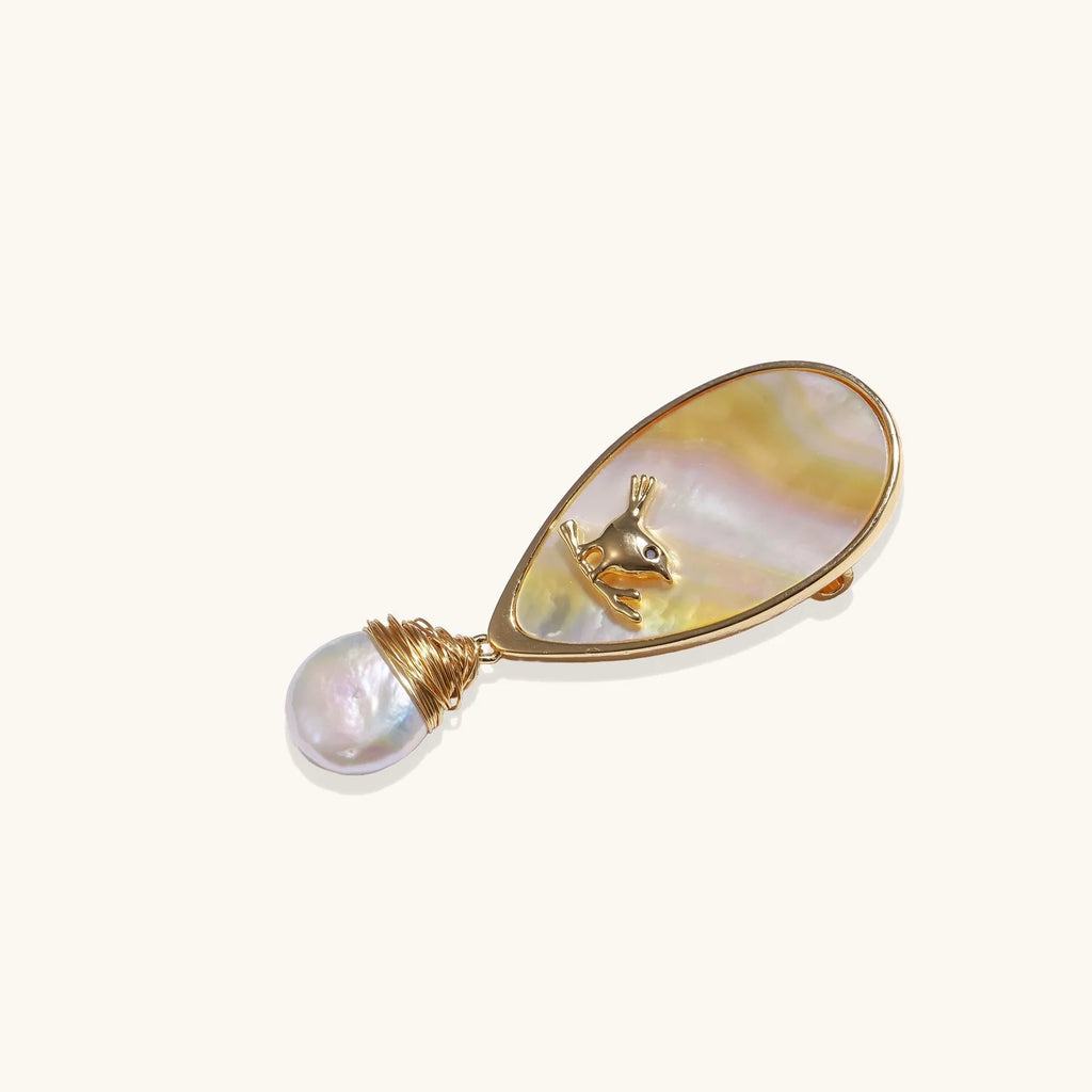 Iridescent shell brooch with gold bird accent and wire-wrapped baroque pearl drop on cream background.