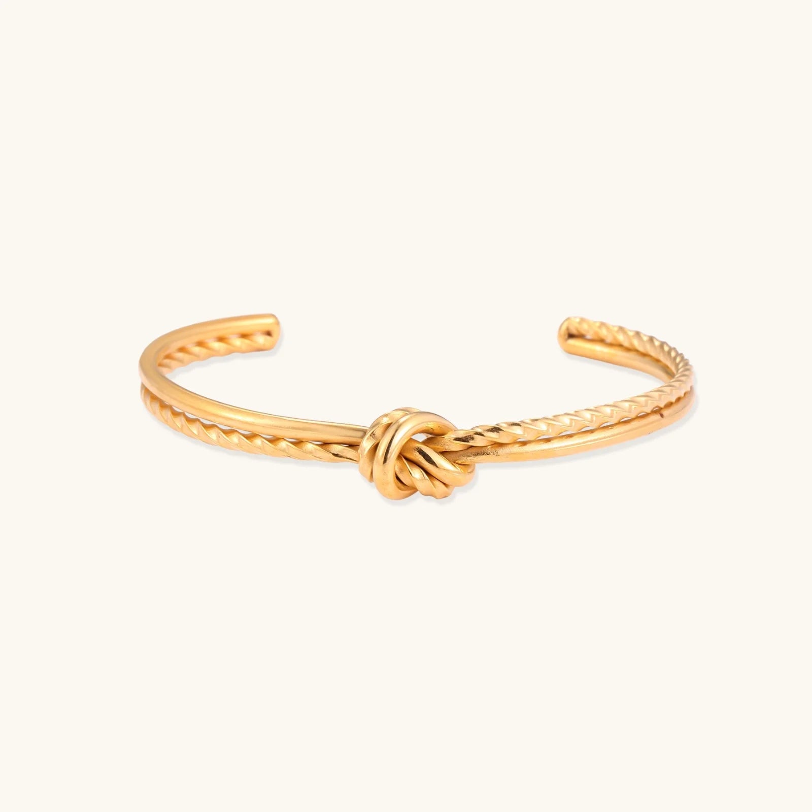 Gold cuff bracelet with twisted rope texture and decorative knot centerpiece on cream background.