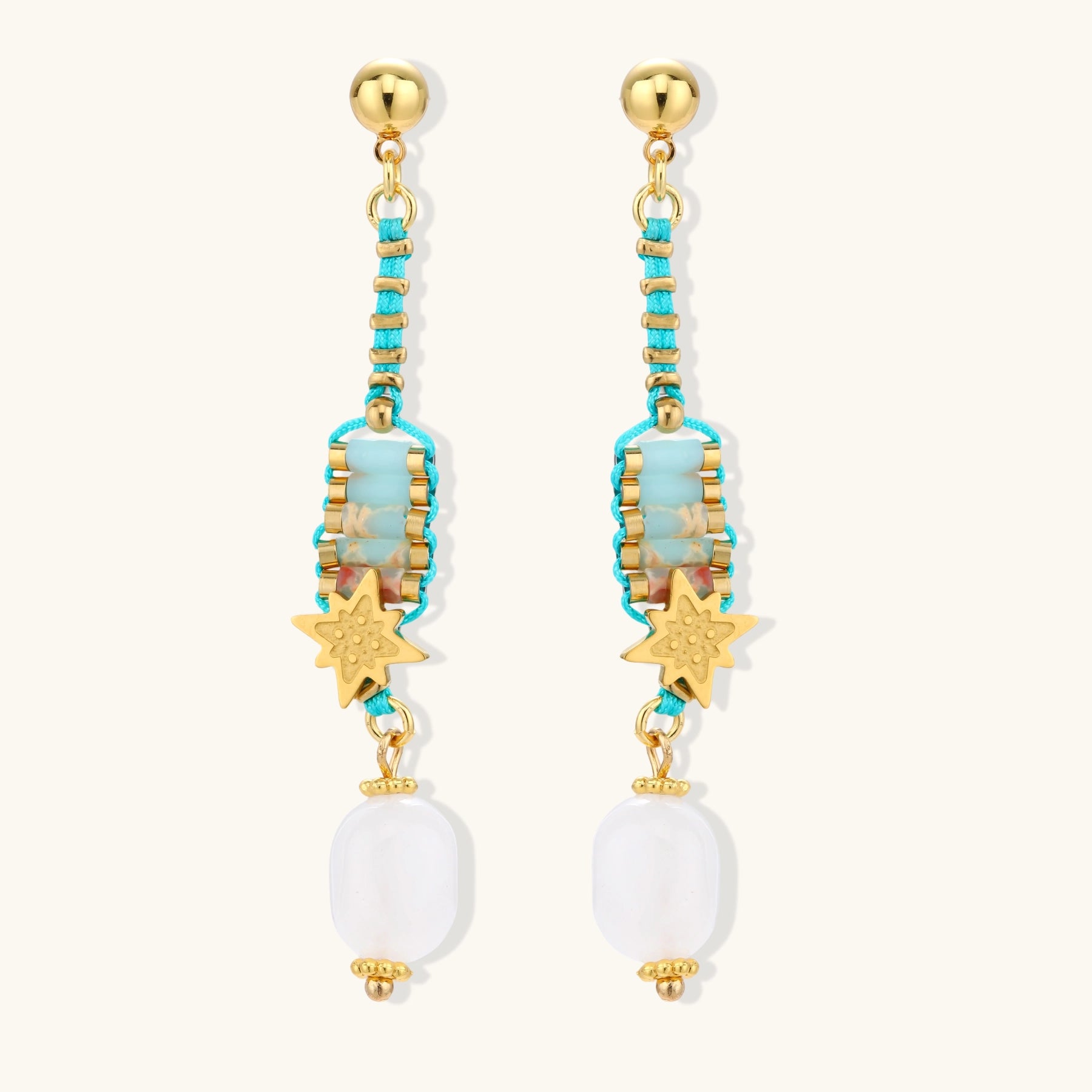 Gold-plated dangle earrings with turquoise threading, star charms, and white pearl beads against cream background.