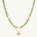 Layered necklace with green stone beads, gold spacers, white accents, and protective eye pendant on cream background.